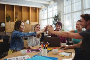 Billedet viser en gruppe kolleger, der samles omkring et bord og giver hinanden high-five i et lyst kontormiljø. Motivet signalerer samarbejde, fælles succes og positiv teamånd i en arbejdssituation.
