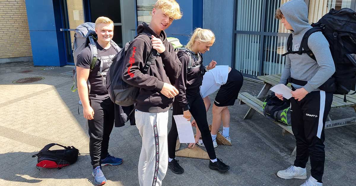 Billedet viser en gruppe unge med rygsække, der er i gang med forberedelser udendørs. Stemningen er afslappet og forventningsfuld, hvor deltagerne gør klar til aktivitet eller fælles start.
