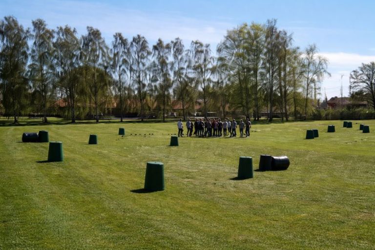 En gruppe unge står samlet på en stor græsplæne, hvor banen er markeret med tønder og forhindringer spredt ud over området. Omgivelserne og opstillingen signalerer en aktiv udendørs teambuilding- eller spilaktivitet med fokus på bevægelse og samarbejde.