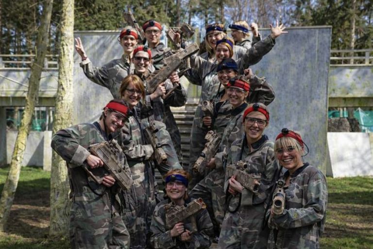 En gruppe deltagere står samlet i camouflagebeklædning med lasergeværer og pandelamper, smilende og med armene i vejret efter en fælles oplevelse. Billedet udstråler holdånd, energi og en afslappet stemning efter en intens og sjov aktivitet.