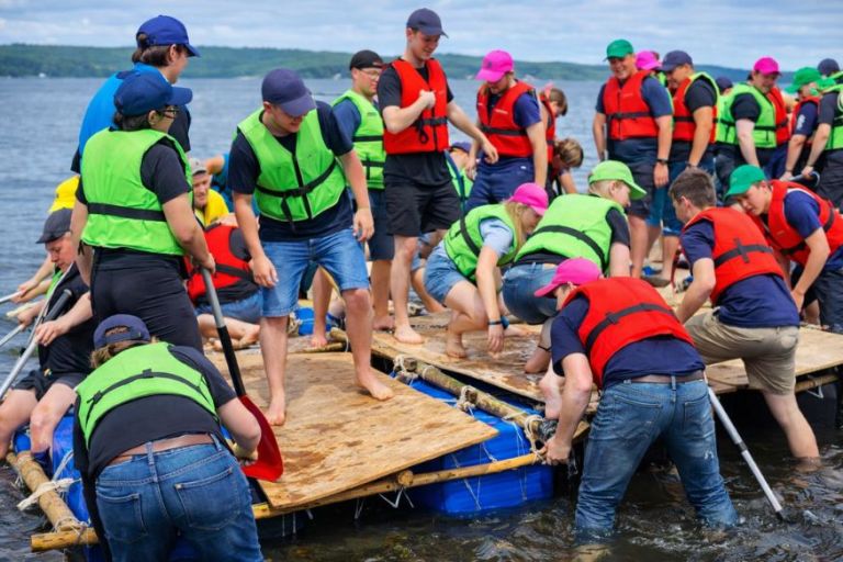 En gruppe unge står tæt sammen på en hjemmelavet tømmerflåde iført redningsveste og kasketter, mens de samarbejder om at holde balancen og komme fremad. Stemningen er præget af grin, koncentration og fælles indsats midt på vandet.