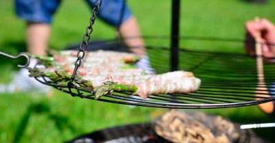 Billedet viser mad, der tilberedes over åben ild på en grillrist udendørs, hvor flere hænder samarbejder om tilberedningen. Motivet signalerer Outdoor Cooking Challenge med fokus på fællesskab, samarbejde og oplevelser i naturen.