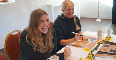 Billedet viser to deltagere, der griner og arbejder koncentreret sammen ved et bord med opgaver og materialer. Motivet udstråler engagement, fællesskab og en afslappet stemning under en samarbejds- eller workshopaktivitet.