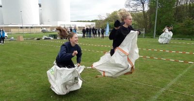 Billedet viser to unge, der deltager i et sækkevæddeløb som en del af en udendørs aktivitet, hvor fart, balance og konkurrence er i fokus. Stemningen er energisk og præget af grin, tilskuere og fælles engagement.