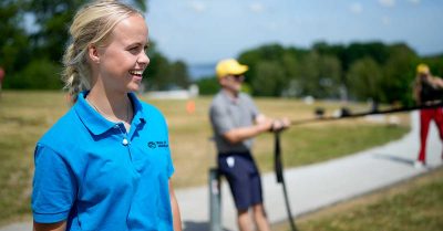 skoleteambuilding-dk-instruktoer1 Billedet viser en smilende instruktør iført blå polo, der observerer en udendørs aktivitet i grønne omgivelser. Motivet signalerer tryghed, overblik og professionel tilstedeværelse under teambuilding og samarbejdsøvelser.