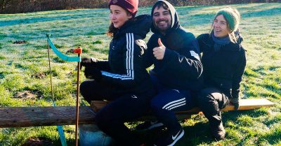 Billedet viser tre deltagere, der sidder tæt sammen på en bænk under en udendørs teambuildingøvelse, hvor samarbejde og balance er i fokus. Stemningen er præget af glæde, fællesskab og succesoplevelse i grønne omgivelser.