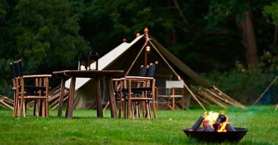 Billedet viser et stemningsfuldt udendørsområde med telt, bålsted og rustikke møbler placeret i grønne omgivelser. Motivet signalerer fællesskab, nærvær og rammer for sociale aktiviteter, teambuilding eller ophold i naturen.
